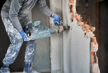 Close up of man builder in workwear drilling wall with hammer drill. Male worker using drill breaker while destroying wall in apartment under renovation. Demolition work and home renovation concept.