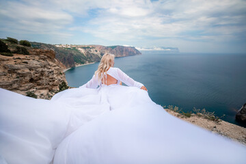 Blonde with long hair on a sunny seashore in a white flowing dress, rear view, silk fabric waving in the wind. Against the backdrop of the blue sky and mountains on the seashore.