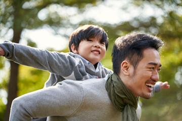 young asian father having fun carrying son on back outdoors in park - obrazy, fototapety, plakaty