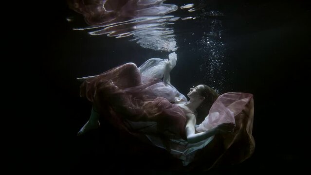 a red-haired woman in translucent fabrics hovers horizontally and beautifully touches hands and feet in a ray of light under the surface of dark water. the general plan
