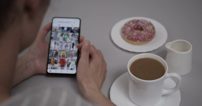 Woman at Home Sitting at Table With Coffee and Donut using Smartphone Scrolling Through Feed. Girl Looks Social Network Page On Mobile Phone. Woman Using Social Network on Smartphone.