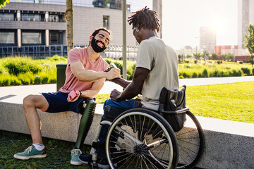 An hispanic man with an artificial prosthesis on his leg shakes hands with his African friend sitting in a wheelchair - diversity lifestyle concept - friendship between people with disabilities - obrazy, fototapety, plakaty
