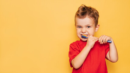 Happy baby boy brushing his teeth with a blue toothbrush on a yellow background. Health care, oral...
