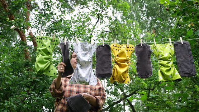 Woman removes dried reusable diapers from clothesline in garden. Modern eco friendly cloth nappy for infant child hygiene. Sustainable lifestyle, zero waste concept. Slow motion.
