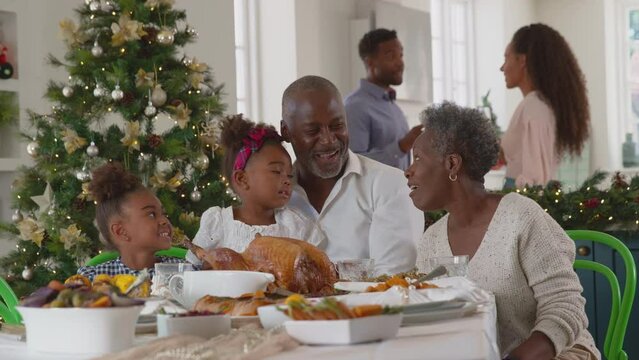 Grandparents sitting at Christmas dinner table with grandchildren as parents prepare meal in background - shot in slow motion
