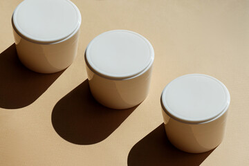White jars of hand cream, faces on beige background in sunlight, with hard shadow. Unmarked...