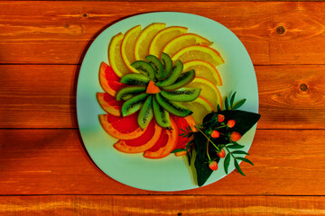 beautifully decorated slices of fresh orange, lemon, banana on a round plate.green kiwi. Lime.Next to the twigs - evergreen pine needles, cones. on a wooden background.the concept of the holiday.	