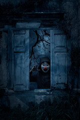 Scary evil clown wear jacket standing in old damaged wood window with wall over dead tree, full...