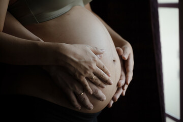 close up of two hands wearing wedding rings on pregnant woman touching her big belly.