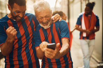 Cheerful senior soccer fan and his son celebrate while looking at results of sports match on smart...