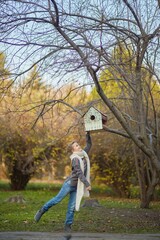 Obraz premium A girl in a knitted sweater in an autumn park looks at a birdhouse. September or October, yellow leaves on trees.