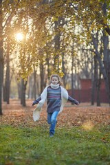 Naklejka na ściany i meble A girl in a knitted sweater in an autumn park runs along the path. September or October, yellow leaves on trees.