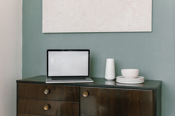 Laptop computer,  ceramics on chest of drawers, blank textured canvas on wall. Blank screen mockup. Styled home office workspace, interior design. Work at home concept.