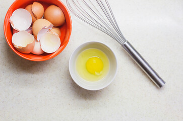 Photography with a highlighted texture on top. The shell of broken eggs, a broken egg in a bowl and a whisk on the white table