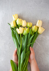 A yound lady arranging a bouquet of yellow tulips on a grey table.