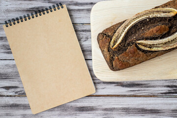 Homemade banana bread on a wooden background with a blank notepad for a recipe or name. Flat lay, top view, copy space