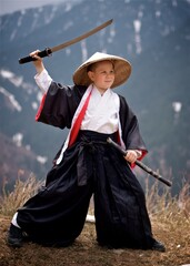 A young caucasian boy seven year old dressed as Samurai in black and white Kimono with katana standing outside in the cloudy spring mountains 