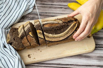 A woman's hand cuts freshly baked homemade banana bread on a wooden background. With raw bananas and a towel on the table.