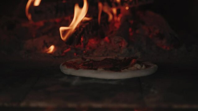 Fresh traditional rustical pizza with salami, green peperoni, cheese and tomatoe souce get shoved into oven fired by wood with beutiful flames at daylight