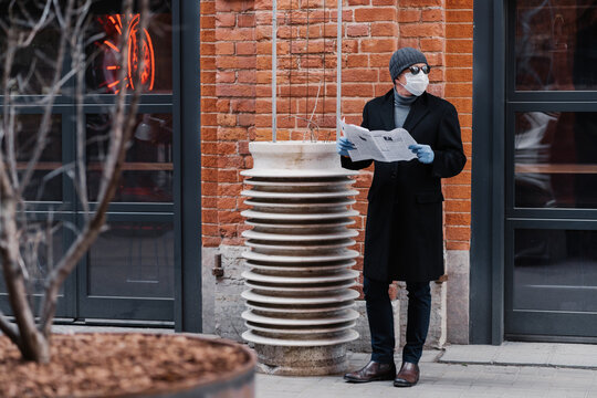 Horizontal shot of young man looks aside, wears medical rubber gloves and face mask, looks aside, holds opened newspaper, reads news, thinks about virus protection. Coronavirus outbreak concept