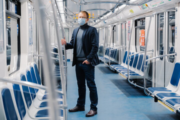 Obraz premium Full length shot of male entrepreneur in formal suit, medical mask, stands in urban train during disease outbreak, virus protection and quarantine protects himself from coronavirus at public transport
