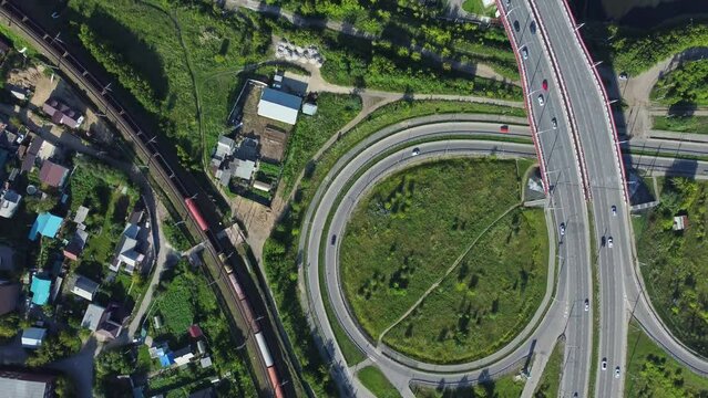 4k Top view of village and cars drive on intersecting roads on summer day irrl. Aerial pic of houses, railroad and traffic driving on winding highway across river in open air. Operator uses drone and