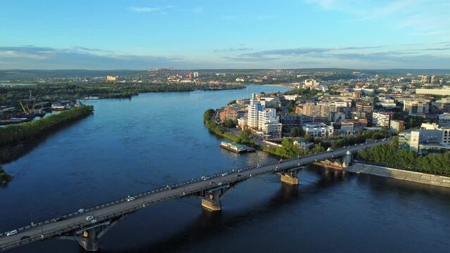 4k Top view of Russia Irkutsk city and bridge with cars on summer day irrl. Aerial pic of town with buildings, river and vehicls driving on road under blue sky. Tourist uses flying drone and shoots