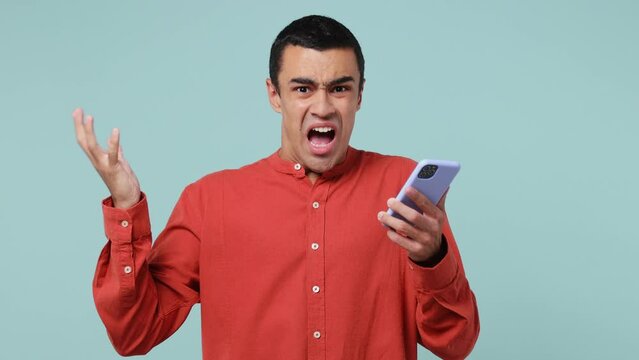 Young sad irritated middle eastern man wear red shirt using mobile cell phone swear spread hands read fake news unexpected rumor has some problem isolated on plain pastel light blue cyan background