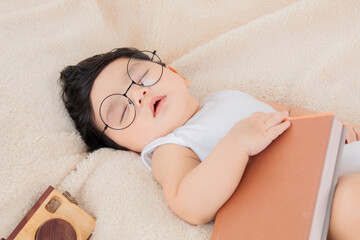 Asian newborn baby wear glasses deeply sleeping and napping with favorite book on beige blanket...
