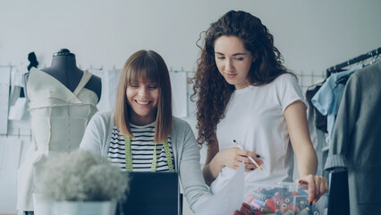 Cheerful creative clothing designers are watching laptop screen, having friendly conversation, gesturing, smiling and laughing together in light workshop. Fun at workplace concept.