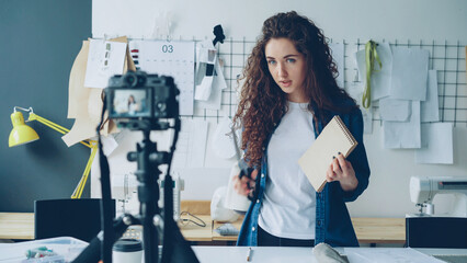 Young attractive seamstress fashion blogger is recording video with camera, talking and showing...