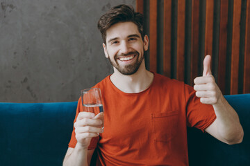 Young man in red t-shirt hold drink clear fresh pure still water from transparent glass show thumb up sit on blue sofa stay at home hotel flat rest relax spend free spare time in living room indoors - obrazy, fototapety, plakaty