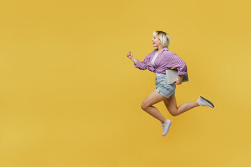 Obraz premium Full bodyside view young blonde IT woman 20s she wear pink tied shirt white t-shirt jump high run fast hurry up hold use work on laptop pc computer isolated on plain yellow background studio portrait.