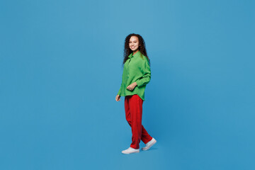 Full body side view cheerful young woman of African American ethnicity 20s she wear green shirt walking going look camera isolated on plain blue background studio portrait. People lifestyle concept.