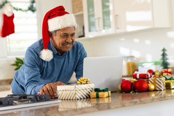 Happy senior biracial man in santa hat making christmas video call on laptop in kitchen