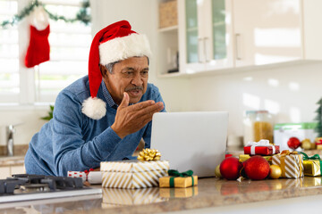 Happy senior biracial man in santa hat making christmas video call on laptop in kitchen