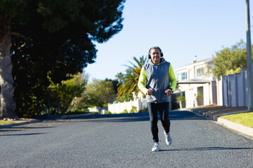 Happy senior biracial man in sports clothes and headphones jogging on sunny road