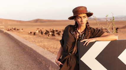 A girl with pigtails in a hat stands near a road sign. In the background is a road in the desert. Morocco.