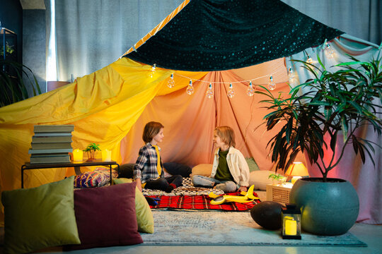 Two young cute boys are playing inside a nice comfy tent that has a hippie vibe to it, the tent is an indoor one