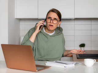 A young woman is sitting in the kitchen in front of a laptop and talking on the phone. On the face of a woman, the emotion of bewilderment of surprise or disappointment