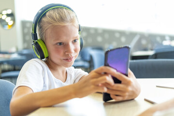 A little girl with headphones is watching cartoons on her phone sitting at a table. Cute schoolgirl listening to music