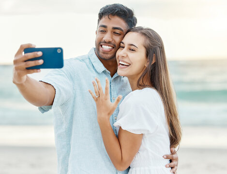 Love, beach and couple after their engagement taking a selfie on a phone while on summer vacation. Happy woman showing off her ring after a proposal while taking picture with a man while on holiday.