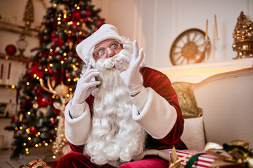 Santa Claus sitting on couch and talking on mobile phone near the fireplace and christmas tree with gifts. New year and Merry Christmas , happy holidays concept