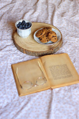 Fototapeta na wymiar Vintage cup with fresh blueberries, plate of chocolate chip cookies, open book and reading glasses on a bed. Selective focus.