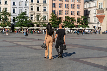 A couple holding hands walking through the old town outdoors on a sunny summer day towards horse-drawn carriages and a summer restaurant in the background, a couple of tourists, adult couple