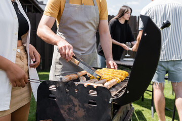 Man in apron cooking on grill near interracial friends in backyard