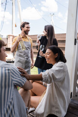 Cheerful bi-racial woman talking to friend near blurred people with wine in backyard
