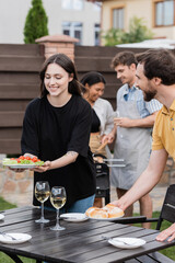 Positive woman holding salad near friend with buns during picnic in backyard