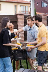 Shocked man in apron putting food on plates near interracial friends and blurred wine on backyard