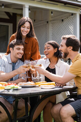Cheerful multiethnic friends toasting with wine near grilled food during picnic outdoors